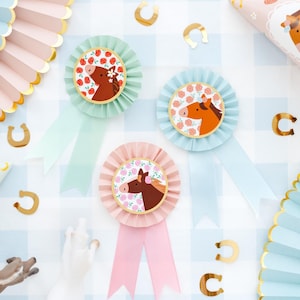 Puede incluir: Tres cintas de roseta con ilustraciones de caballos y estampados florales. Las rosetas son en verde pastel, azul y rosa, cada una con una cinta a juego. Confeti de herraduras doradas y abanicos de papel decorativos completan el tema ecuestre.
