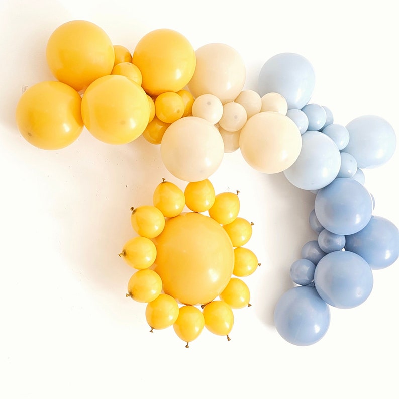May include: A balloon arch with yellow, cream, and blue balloons. The arch is shaped like a sun with a large yellow balloon in the center.