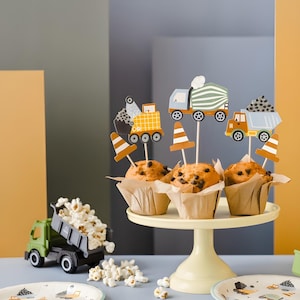 Puede incluir: Una presentación festiva con magdalenas con chispas de chocolate decoradas con adornos temáticos de construcción, incluyendo camiones y conos de tráfico. Un camión volquete de juguete derrama palomitas de maíz. El conjunto está sobre un soporte para pasteles.