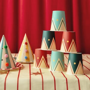Puede incluir: Una colección de vasos de papel y gorros de fiesta cónicos. Los vasos están apilados en pirámide y presentan patrones geométricos de montañas en rojo, verde oscuro y azul claro con puntos amarillos. Dos gorros de fiesta tienen pompones de colores.
