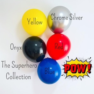 Puede incluir: Cinco globos de diferentes colores: amarillo, plata cromado, rojo, negro y azul. Los globos están dispuestos en círculo con el texto "The Superhero Collection" y "POW!" en el centro.