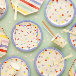 Puede incluir: Vista aérea de suministros para fiestas sobre una superficie verde claro. Incluye platos con bordes azules y lunares coloridos, platos a rayas, vasos de papel, servilletas, tenedores y pajitas. Confeti esparcido alrededor.
