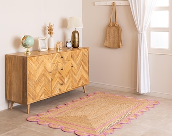 May include: A rectangular jute rug with a scalloped pink border. The rug is on a light-colored tile floor. A wooden cabinet with a herringbone pattern is in the background, along with a globe, lamp, and clock.