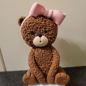 Könnte beinhalten: Eine braune Teddybär-Figur mit einer rosa Schleife auf dem Kopf. Der Bär hat eine strukturierte Oberfläche und sitzt auf einem weißen Sockel. Nase und Mund des Bären sind hellbeige.