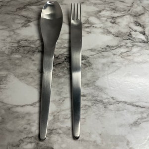 May include: A set of silver cutlery, including a spoon and a fork, on a white marble background.