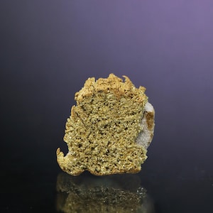 May include: A close-up of a gold nugget with a rough, textured surface. The nugget is a light gold color and has a white, powdery coating on one side.