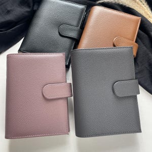 Könnte beinhalten: Vier ledergebundene Planer in Schwarz, Braun, Grau und Mauve. Jeder Planer hat eine strukturierte Oberfläche und einen gebogenen Laschenverschluss. Die Planer sind auf einer weißen Oberfläche mit schwarzem Stoff und getrockneten Pflanzen im Hintergrund angeordnet.
