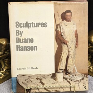 May include: A book titled "Sculptures By Duane Hanson" with a photo of a man in overalls holding a paint roller on the cover. The book is by Martin H. Bush.
