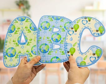 May include: Three large cardboard letters, A, B, and C, are decorated with a blue and green pattern of the Earth with a smiling face. The letters are held up by two hands.