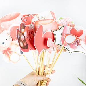 May include: A set of Valentine's Day themed photo booth props. The props are made of paper and feature various designs, including hearts, teddy bears, and glasses. The props are attached to wooden sticks.