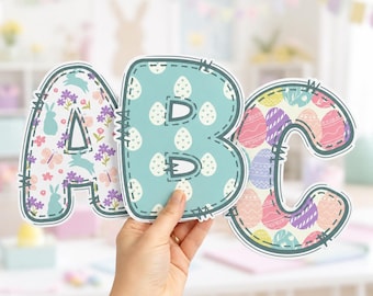 May include: Three large cutout letters A, B, and C featuring decorative patterns. The letter A has a floral and bunny print, the letter B has a white egg pattern on a teal background, and the letter C features a colorful Easter egg design.