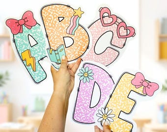May include: Colorful, decorative letters A, B, C, D, and E. Each letter has a unique design, including a bow, sunglasses, a lightning bolt, a book, a star, a rainbow, and daisies. The letters are held up by a person.