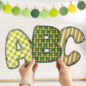 May include: Three large felt letters, A, B, and C, in green, yellow, and orange, with a shamrock and horseshoe pattern. The letters are being held by hands.