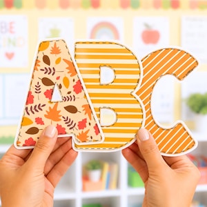 Op de afbeelding: De letters A, B en C worden voor een klasachtergrond gehouden. De letter A heeft een herfstbladerenpatroon. De letter B heeft horizontale strepen en de letter C heeft diagonale strepen. De letters zijn oranje, wit en bruin.