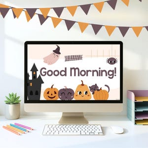 Könnte beinhalten: Ein Computermonitor zeigt eine Halloween-Grafik mit dem Text "Good Morning!" und Cartoon-Kürbissen, einem Geist und einer Spinne. Eine Girlande aus orangefarbenen und lila Wimpeln hängt über dem Monitor. Eine Tastatur, eine Maus und Bleistifte befinden sich auf dem weißen Schreibtisch.