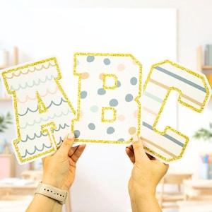 Pode incluir: Três letras grandes de cartão que soletram "ABC" com um contorno de glitter dourado. Cada letra tem um padrão diferente: ondas, bolinhas e listras. As letras são seguradas por duas mãos.