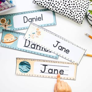 Pode incluir: Dois crachás de nome imprimíveis com um espaço para uma criança escrever seu nome. Os crachás de nome apresentam um tema espacial azul e branco com planetas e estrelas. O primeiro crachá de nome diz "Daniel" e o segundo crachá de nome diz "Jane".