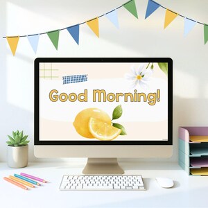 May include: A desktop computer displays a "Good Morning!" graphic featuring a lemon illustration. The setup includes a keyboard, mouse, and colorful pencils on a white desk. A succulent plant in a pot and a multi-tiered paper organizer are also visible.