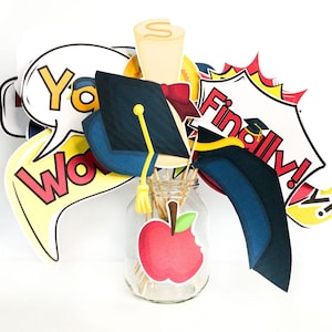 Puede incluir: Un conjunto de accesorios para cabina de fotos de graduación con un gorro de graduación, un diploma, una manzana y burbujas de diálogo con las palabras "¡Sí!" "¡Guau!" y "¡Por fin!"