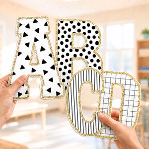 Può includere: Quattro grandi lettere fustellate A, B, C e D con distinti motivi in bianco e nero. La lettera A presenta triangoli neri, B pois neri, C strisce verticali nere e D una griglia nera, il tutto bordato con glitter color oro.
