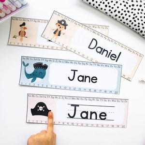May include: Three printable name tags with a nautical theme. The first tag features a blue whale with a pirate hat, the second tag features a pirate hat with a skull and crossbones, and the third tag has a blank line for writing a name. Each tag has a number line with numbers 0-20.