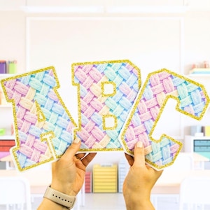 May include: Three large, colorful letters spelling "ABC" are held up. The letters have a woven pattern in pastel colors and are outlined with gold glitter. The background is a classroom setting.
