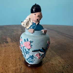 Puede incluir: Pequeña figura decorativa. Una figura con un atuendo azul y un sombrero negro emerge de un jarrón azul claro. El jarrón está adornado con diseños florales rosas y hojas azules. Un perro blanco asoma por encima del jarrón.