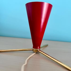 Puede incluir: Una lámpara roja en forma de cono con una base de trípode de latón. La lámpara tiene un acabado brillante y un cable de alimentación blanco. La base tiene tres patas de latón. La lámpara está sobre una superficie clara con un fondo azul.