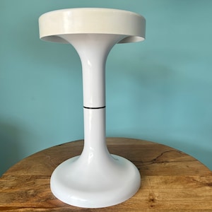 Könnte beinhalten: Weiße Tischlampe mit einem breiten, runden Sockel und einem hohen, schlanken Schaft. Die Lampe hat einen weißen Schirm mit einem schwarzen Band um den Schaft.