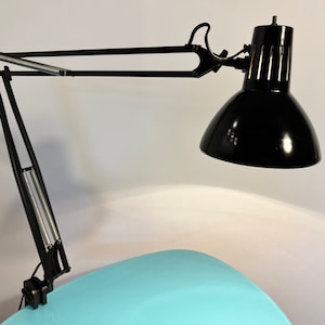 Puede incluir: Lámpara de escritorio negra ajustable con una pantalla negra y un brazo plateado. La lámpara está unida a una superficie azul.