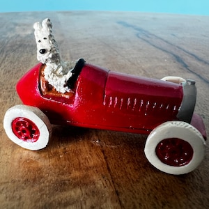 Puede incluir: Un coche de carreras de juguete rojo con ruedas blancas y una figura de animal blanco y esponjoso en el asiento del conductor. El coche tiene un acabado brillante y está sobre una superficie de madera y un fondo azul.