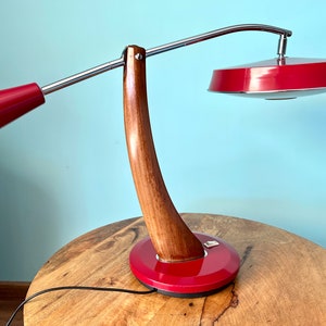 May include: A red and wood desk lamp with a curved wooden base and a chrome arm. The lamp has two red shades, one round and one cone-shaped.