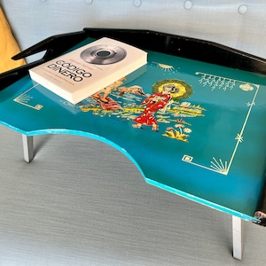 Puede incluir: Una mesa de bandeja azul con ribete negro y patas plateadas. La bandeja presenta una ilustración colorida de una mujer con un kimono rojo y un diseño floral. La mesa de bandeja tiene un libro encima con el título "CODIGO DINERO".