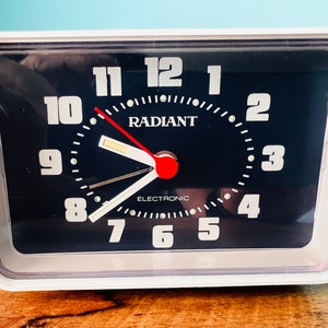 Puede incluir: Un despertador de plástico blanco con una esfera negra. La esfera del reloj tiene números y manecillas plateadas, y la palabra "RADIANT" en el centro. El reloj también dice "ELECTRONIC" debajo de la palabra "RADIANT".