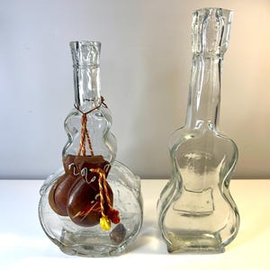 Puede incluir: Dos botellas de vidrio transparente con forma de instrumentos musicales. La botella de la izquierda tiene forma de guitarra y contiene dos pequeñas castañuelas de cuero marrón en su interior. La botella de la derecha tiene forma de violín.