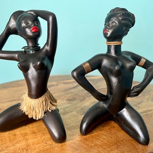 Puede incluir: Dos figuritas de cerámica negra de mujeres con labios rojos. Una lleva una falda de hierba marrón y la otra lleva brazaletes dorados. Ambas están arrodilladas con las manos en las caderas.