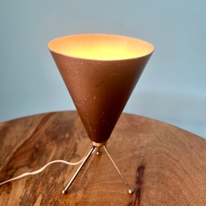 Może przedstawiać: Nowoczesna lampa stołowa z połowy wieku z kloszem w kształcie stożka w kolorze miedzi i podstawą z trzema nogami wykonaną z pozłacanego metalu.