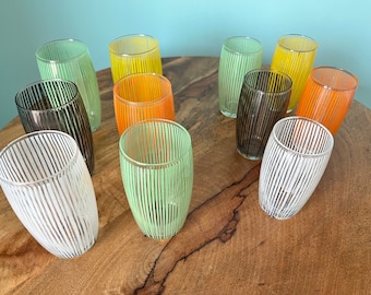 Pode incluir: Uma coleção de copos de beber listrados em várias cores, incluindo verde, amarelo, laranja e preto. Os copos estão dispostos sobre uma superfície de madeira.