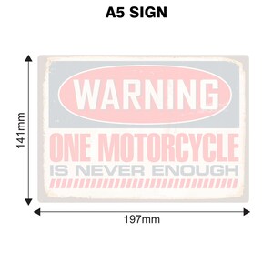 May include: A white sign with a pink oval that says "WARNING" in white text. The sign also says "ONE MOTORCYCLE IS NEVER ENOUGH" in black text. The sign is 197 mm wide and 141 mm tall.