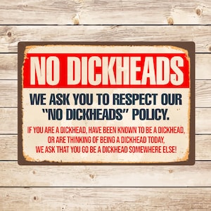 Op de afbeelding: Een rechthoekig metalen bord met een bruine rand en een houten achtergrond. Het bord heeft de woorden "NO DICKHEADS" in grote rode letters bovenaan. Daaronder staat "WE ASK YOU TO RESPECT OUR 'NO DICKHEADS' POLICY."