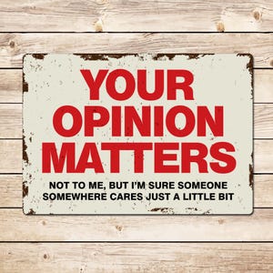 Peut inclure: Une plaque en métal blanc avec un aspect vieilli et rouillé. La plaque porte un texte rouge qui dit "YOUR OPINION MATTERS" et un texte noir plus petit qui dit "NOT TO ME, BUT I'M SURE SOMEONE SOMEWHERE CARES JUST A LITTLE BIT".