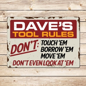 May include: A rectangular metal sign with a distressed finish. The sign features the text "DAVE'S TOOL RULES" in bold white and yellow letters on a maroon background. Below, the word "DON'T" is in red, followed by rules in black.