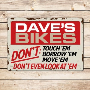 Peut inclure: Une plaque en métal rouillée avec le texte "DAVE'S BIKES" en lettres rouges et blanches. La plaque comporte également le texte "DON'T TOUCH 'EM BORROW 'EM MOVE 'EM DON'T EVEN LOOK AT 'EM" en lettres rouges.