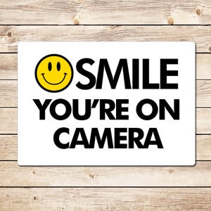 May include: White sign with black text that reads "SMILE YOU'RE ON CAMERA" with a yellow smiley face icon.