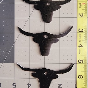 May include: Three black leather cowhide patches in the shape of a bull's head with holes in the center. The patches are on a gray grid background.