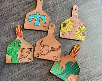 May include: Five leather keychains shaped like cattle ear tags. Each tag features a different design: a turquoise bird, a skull, a cactus, sunflowers, and a cactus with a star. The tags are all brown with a light brown border and the text "MK" on the bottom.