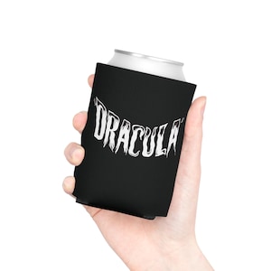 May include: A black foam can cooler sleeve featuring the word DRACULA printed in a white, stylized, dripping font across the center. The sleeve is designed to fit around a standard beverage can to provide insulation and a comfortable grip.