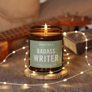 May include: A brown glass candle with a white label that reads "Smells Like A Badass Writer". The candle is lit and sitting on a wooden coaster. The candle is surrounded by white string lights.