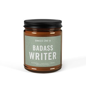May include: A brown glass candle jar with a black lid. The label reads "Smells Like A Badass Writer" with the text "Ecofriendly / Non-Toxic / Vegan 200funnygiftideas" below.