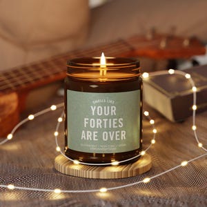 May include: A brown glass candle with a white label that reads "Smells Like Your Forties Are Over". The candle is lit and sitting on a wooden coaster.